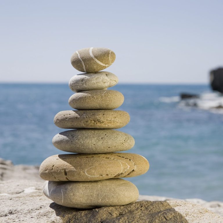Systemisches Coaching und Persönlichkeitsentwicklung Ein Turm aus neun sanft geschliffenen Steinen am Küstenrand, mit Meer im Hintergrund.